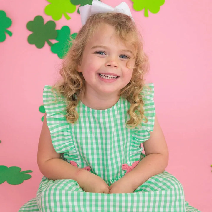 Young girl in a green checkered dress with a white bow sitting on a pink background with shamrock decorations.