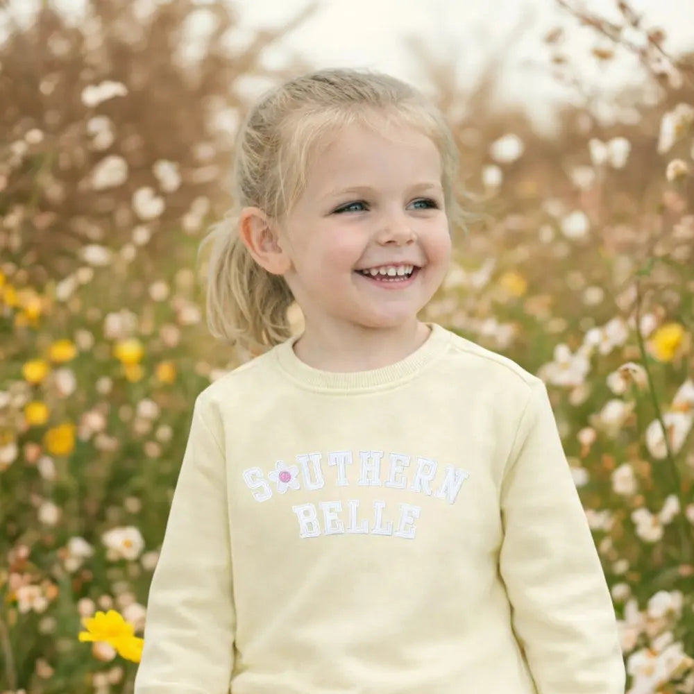 Young girl in a yellow outfit with 'Southern Belle' text, standing in a field of flowers.