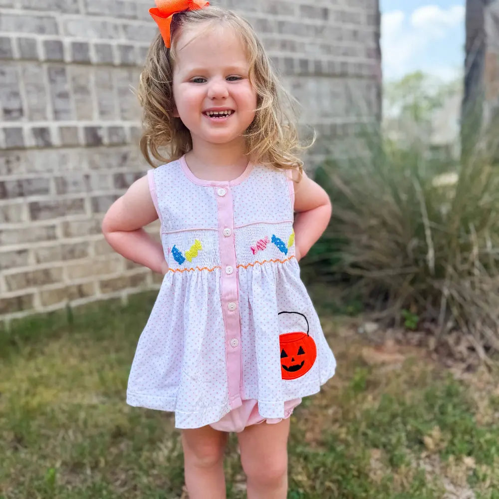 Young girl in a white dress with colorful accents standing outdoors against a brick wall.