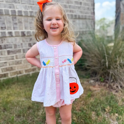 Young girl in a white dress with colorful accents standing outdoors against a brick wall.