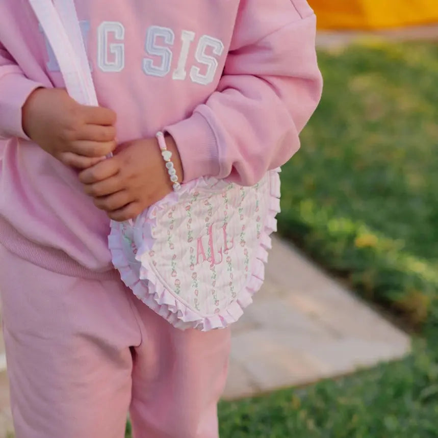 Person wearing a pink 'BIG SIS' sweatshirt holding a heart-shaped pillow with 'LIL' on it, outdoors.