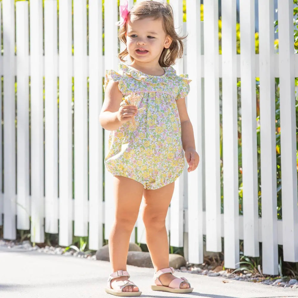 Child wearing a floral romper standing in front of a white picket fence.