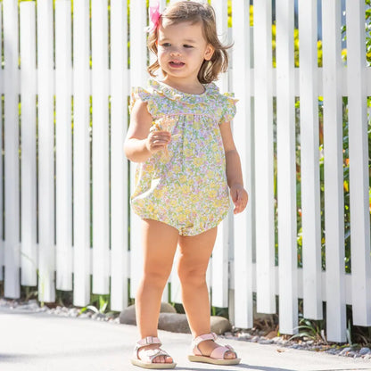 Child wearing a floral romper standing in front of a white picket fence.