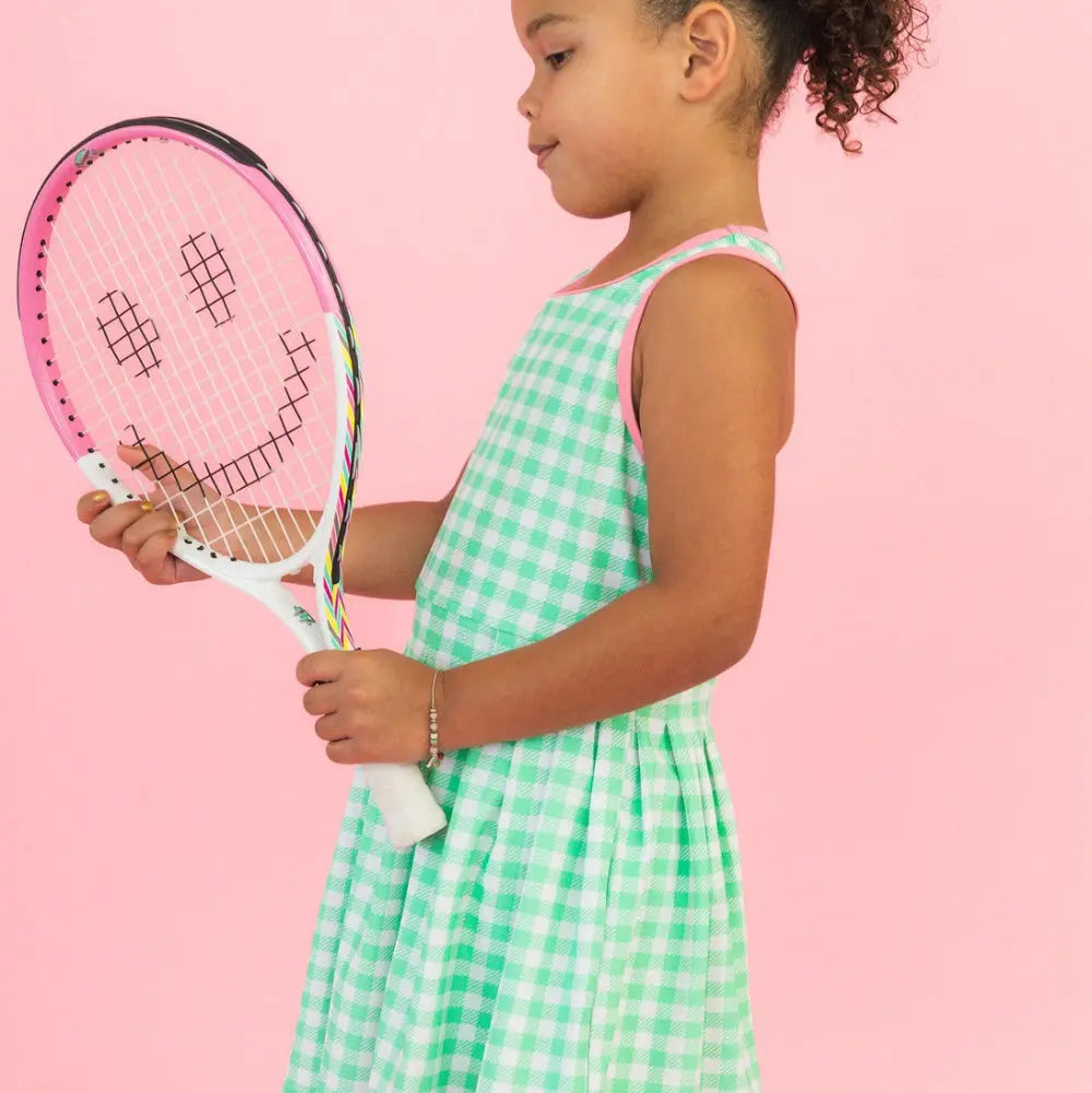 Young girl in a green checkered dress holding a pink tennis racket with a smiley face on a pink background