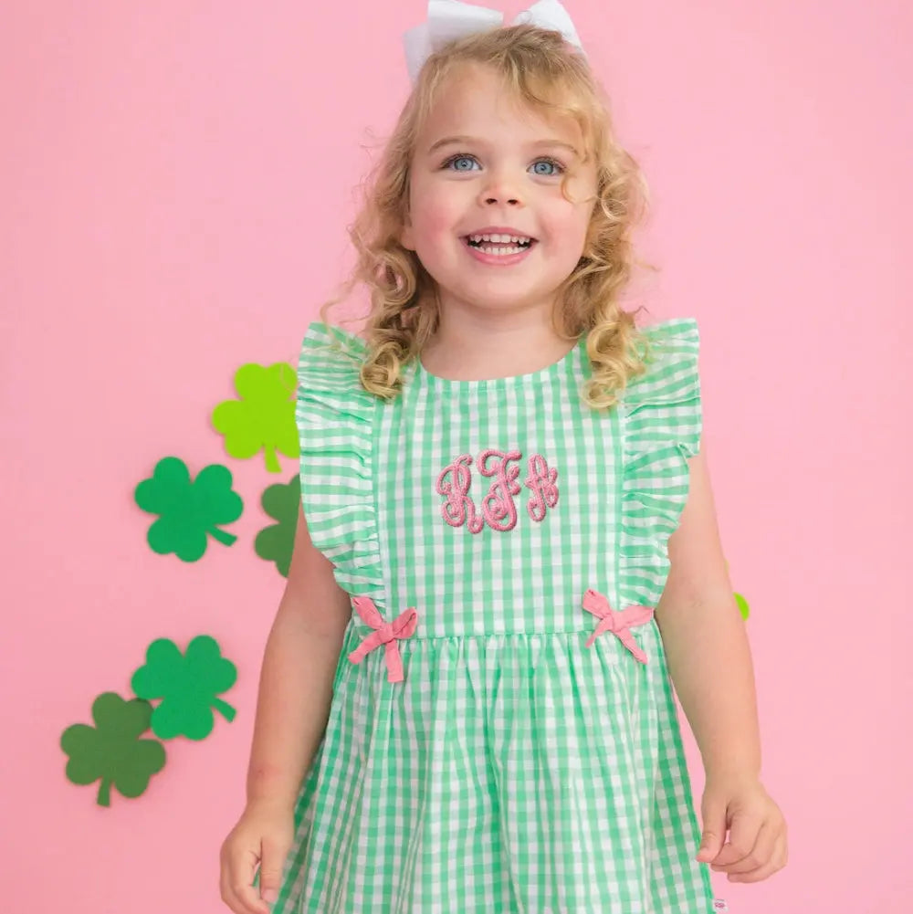 Young girl wearing a green checkered dress with pink bows and monogram on a pink background with shamrock decorations.