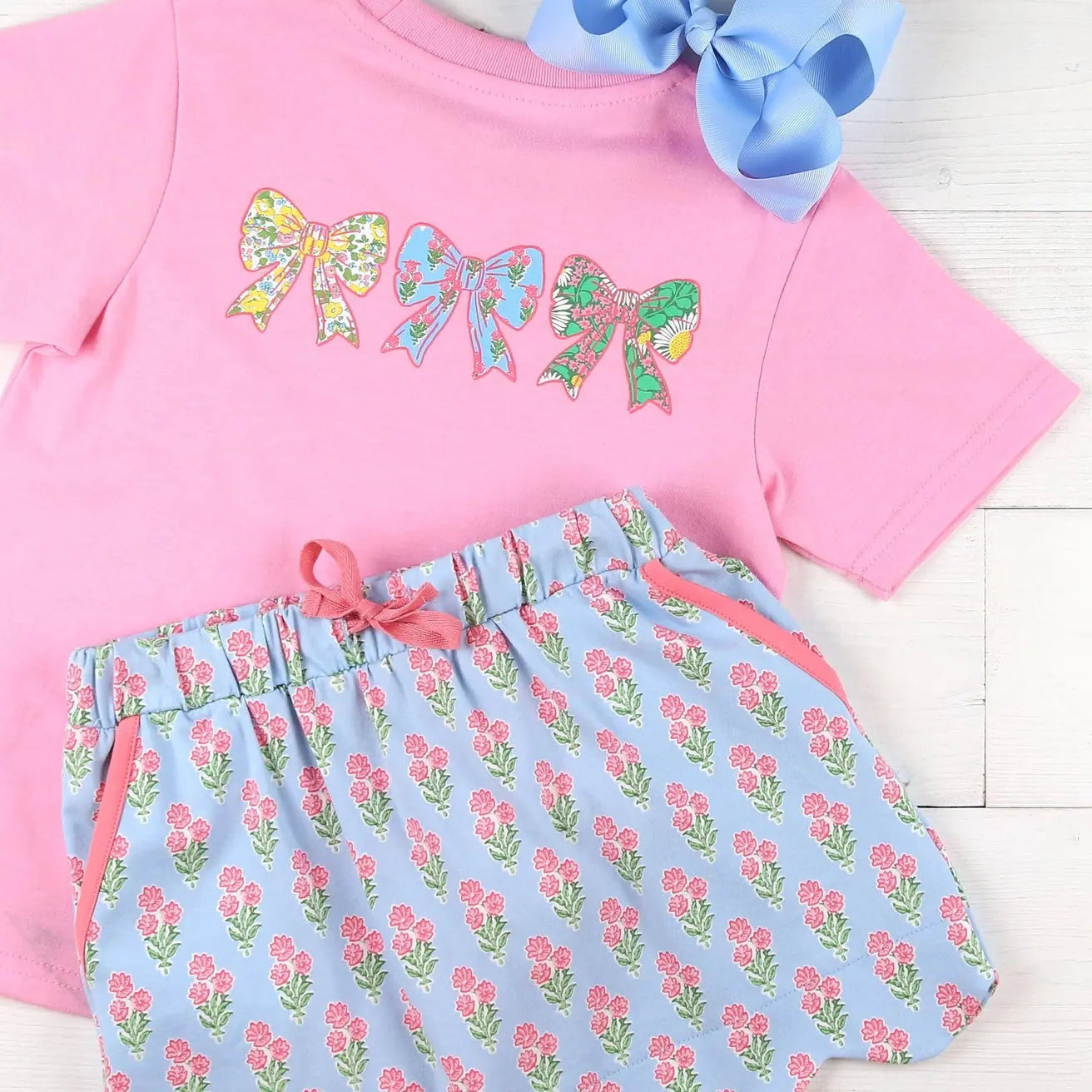 Pink shirt with colorful bow designs and floral shorts on a white background