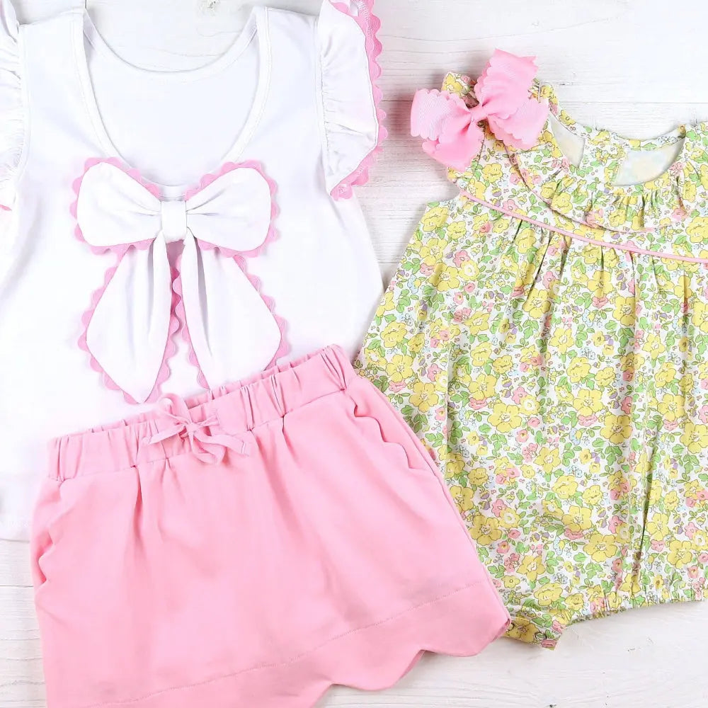 Children's outfit with white top, pink shorts, and floral dress on a light wooden surface.