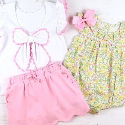 Children's outfit with white top, pink shorts, and floral dress on a light wooden surface.