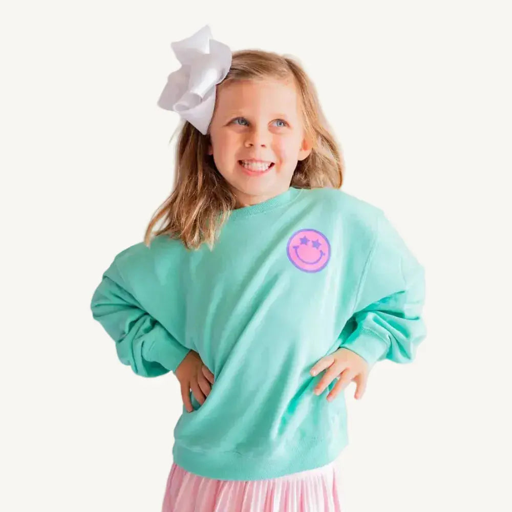 Young girl wearing a teal sweatshirt with a pink logo and a pink skirt, standing against a plain background.