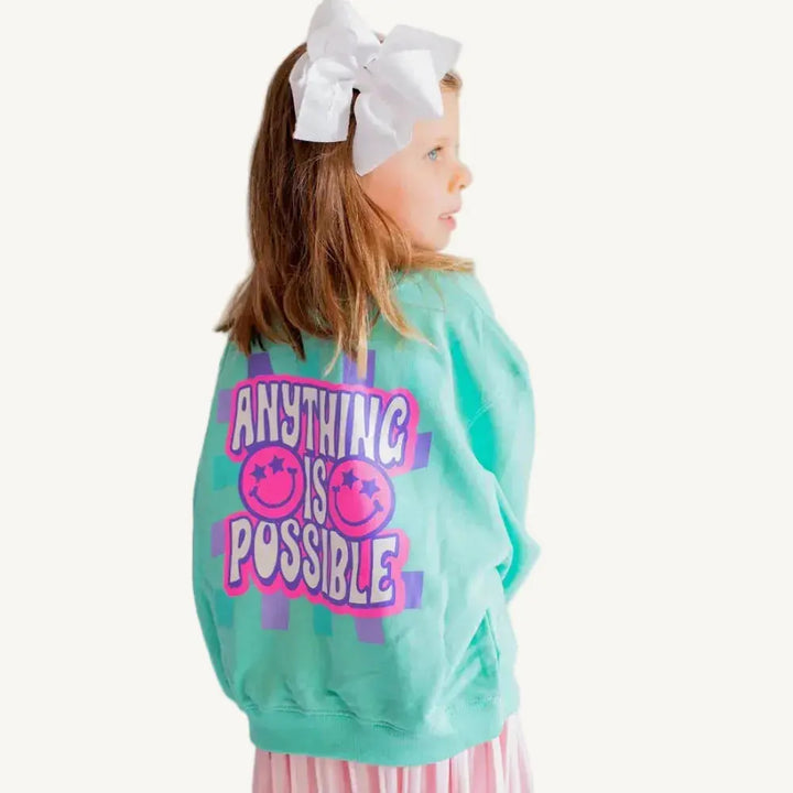 Young girl wearing a teal sweatshirt with 'Anything is Possible' text and a white bow in her hair.