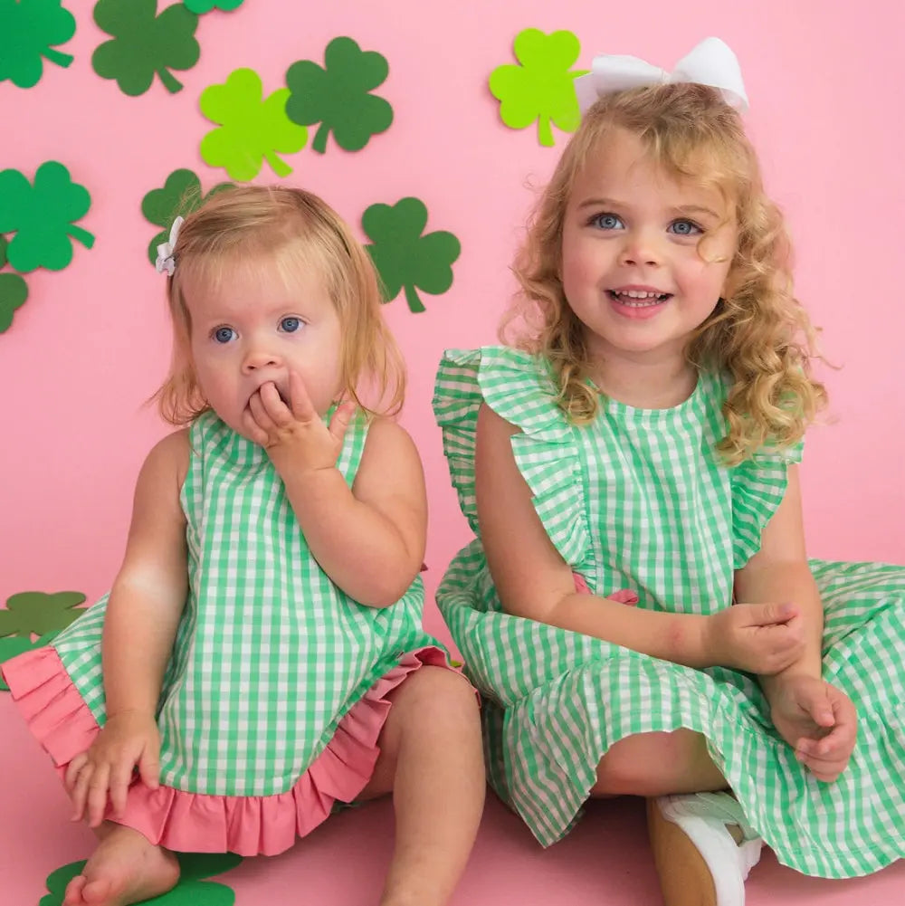 Two young girls in green and pink checkered dresses sitting on a pink background with green shamrock decorations.
