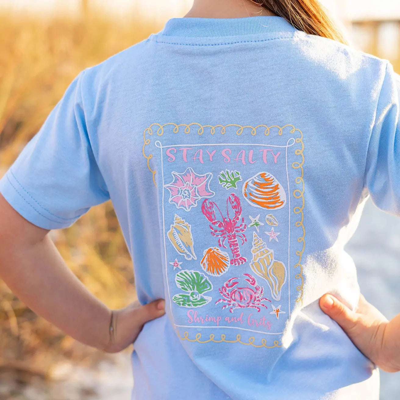 Light blue t-shirt with 'Stay Salty' design featuring sea creatures, worn by a person outdoors.