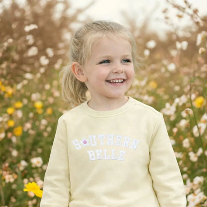 Young girl in a yellow outfit with 'Southern Belle' text, standing in a field of flowers.